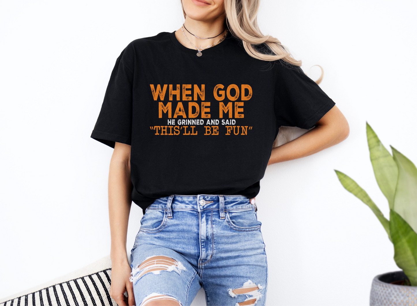 Woman wearing black unisex softstyle t-shirt with orange text "When God Made Me He Grinned and Said 'This'll Be Fun'"