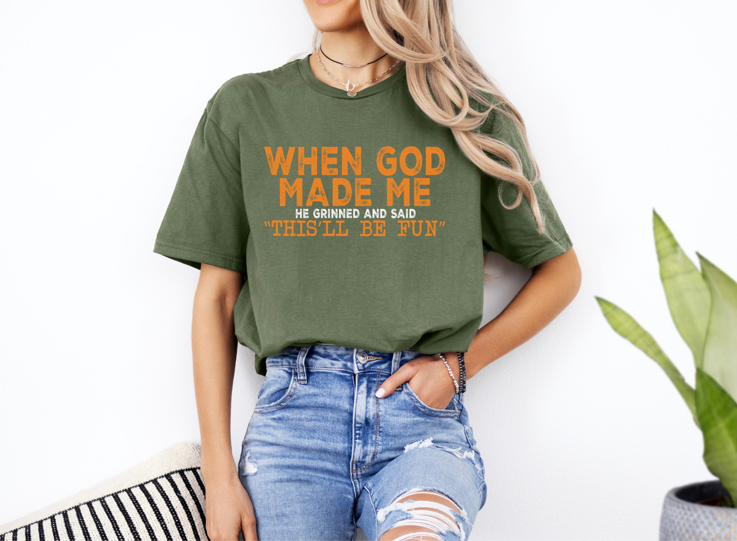 Unisex softstyle green t-shirt with orange text "WHEN GOD MADE ME He grinned and said 'THIS'LL BE FUN'" worn with ripped blue jeans.
