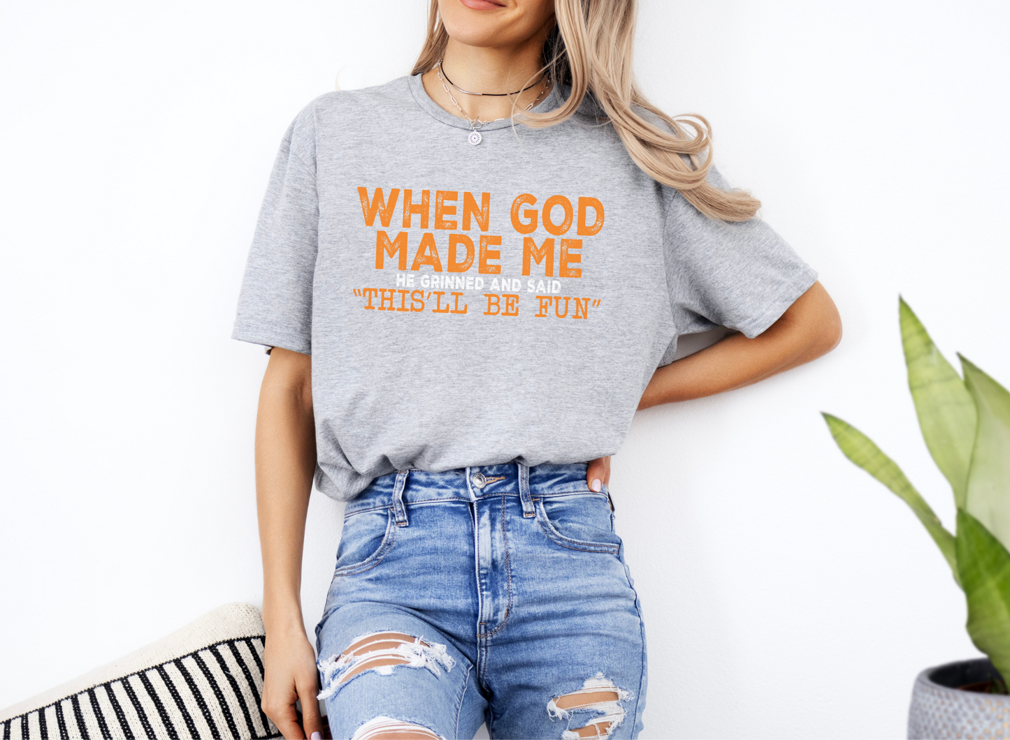 Unisex softstyle grey t-shirt with orange text "When God Made Me He Grinned and Said 'This'll Be Fun'" paired with ripped blue jeans.