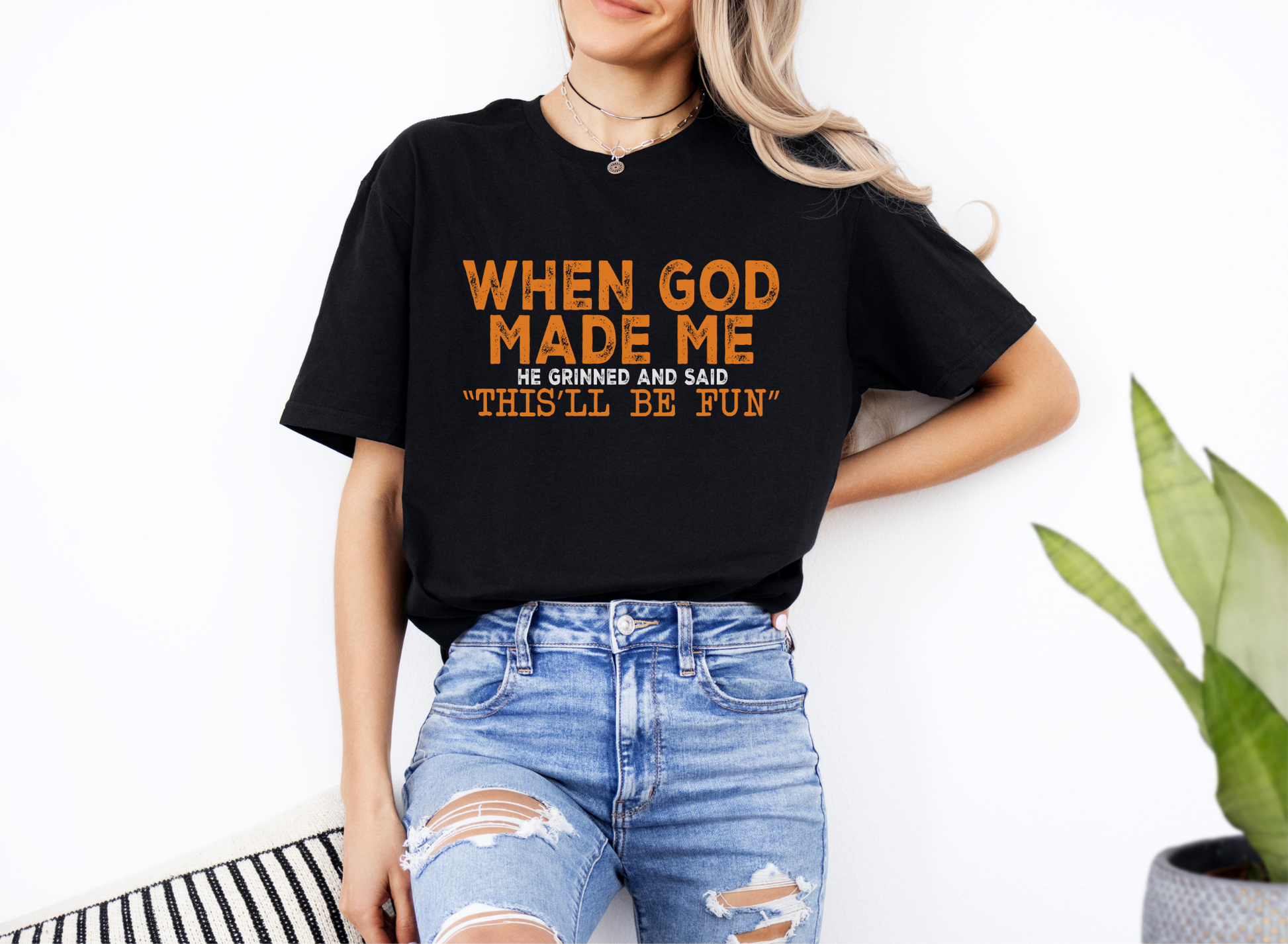Woman wearing black unisex softstyle t-shirt with orange text "When God Made Me He Grinned and Said 'This'll Be Fun'"