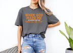 Woman wearing gray softstyle t-shirt with orange text "When God Made Me He Grinned and Said 'This'll Be Fun'" and ripped blue jeans.