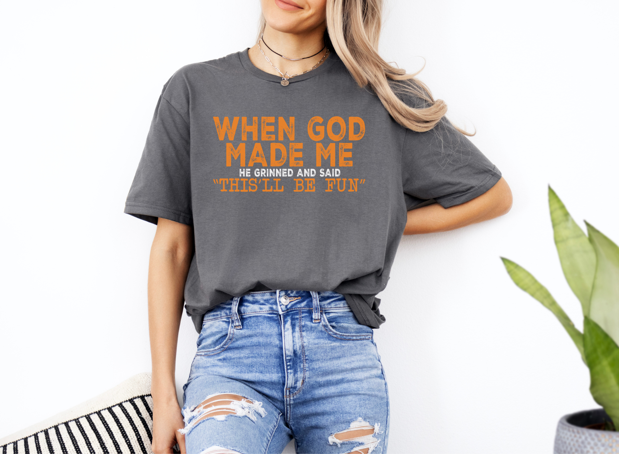 Woman wearing gray softstyle t-shirt with orange text "When God Made Me He Grinned and Said 'This'll Be Fun'" and ripped blue jeans.