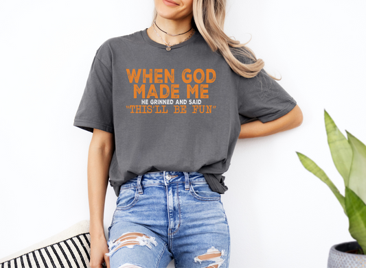 Woman wearing gray softstyle t-shirt with orange text "When God Made Me He Grinned and Said 'This'll Be Fun'" and ripped blue jeans.
