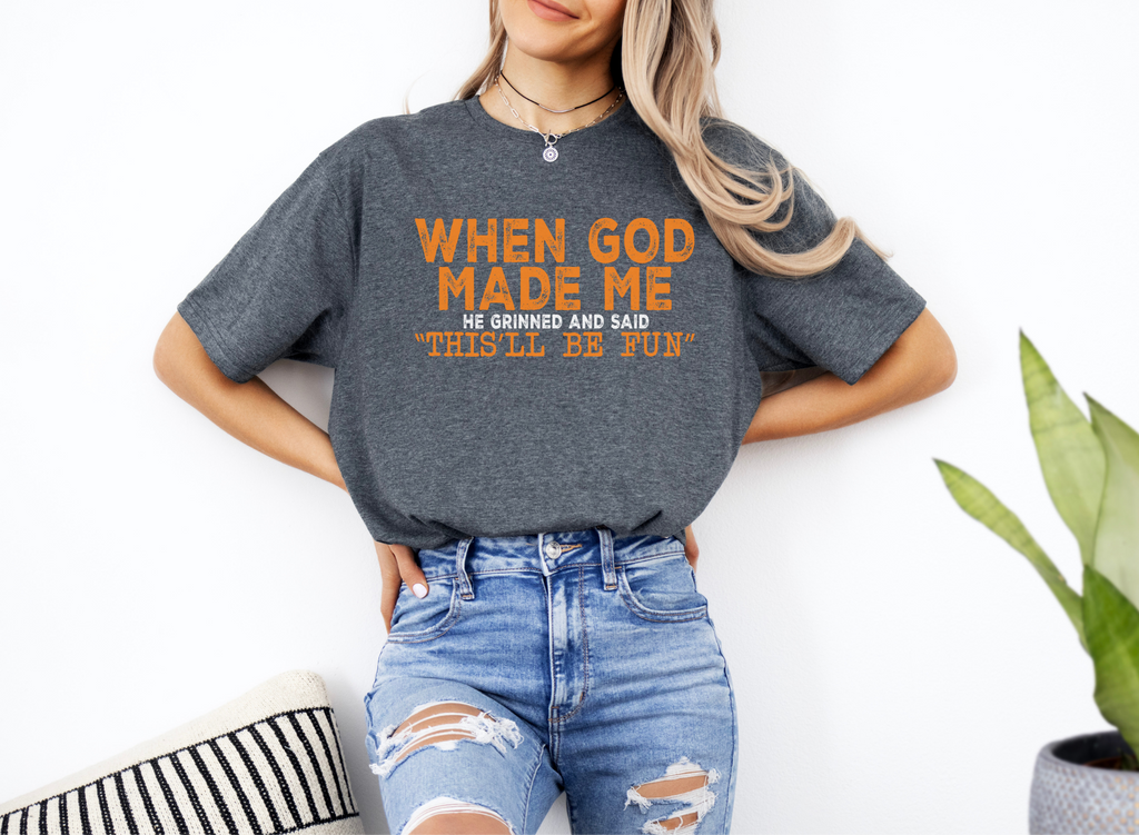 Woman wearing a dark gray unisex softstyle t-shirt with orange text "When God Made Me He Grinned and Said This'll Be Fun" paired with ripped blue jeans.