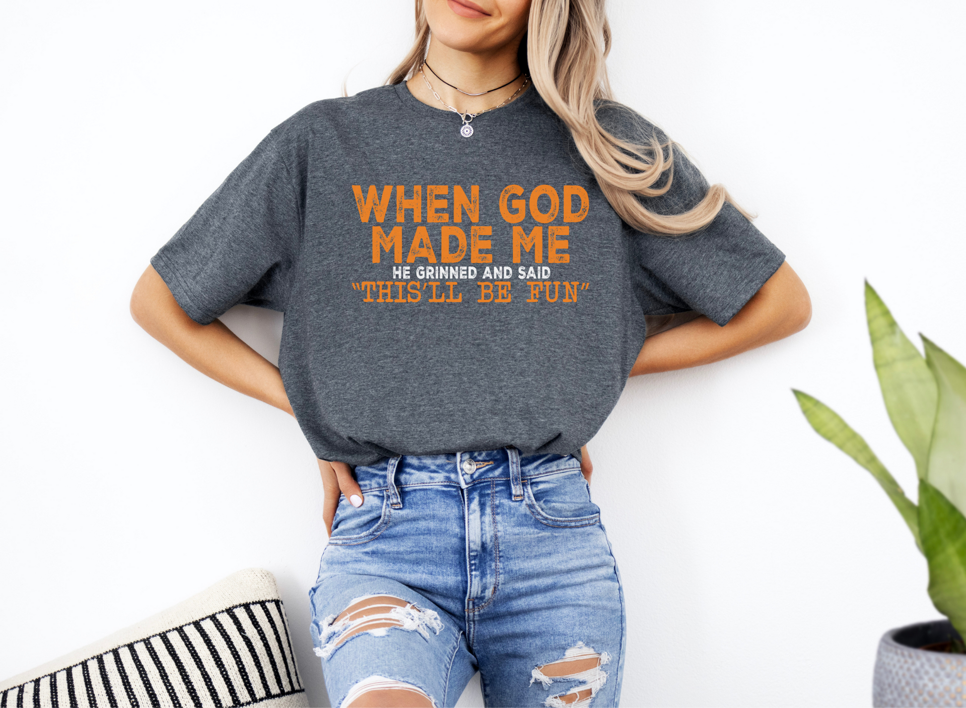 Woman wearing a dark gray unisex softstyle t-shirt with orange text "When God Made Me He Grinned and Said This'll Be Fun" paired with ripped blue jeans.