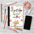 Motivational journal with pink floral cover and personalized name on wooden desk with pens, phone, earphones, and scrunchie.