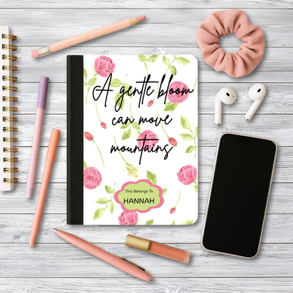 Motivational journal with pink floral cover and personalized name on wooden desk with pens, phone, earphones, and scrunchie.