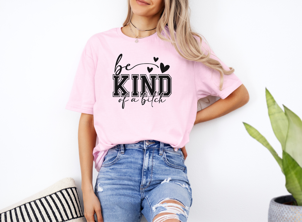 Woman wearing pink softstyle t-shirt with bold "Be Kind of a Bitch" text, paired with ripped blue jeans.