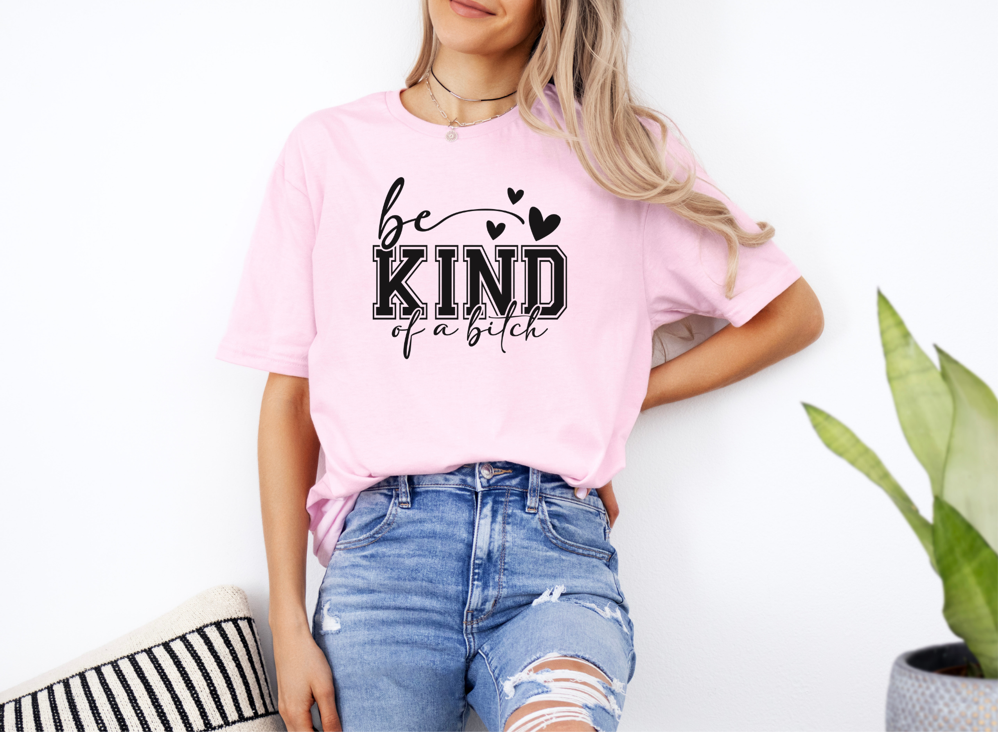 Woman wearing pink softstyle t-shirt with bold "Be Kind of a Bitch" text, paired with ripped blue jeans.