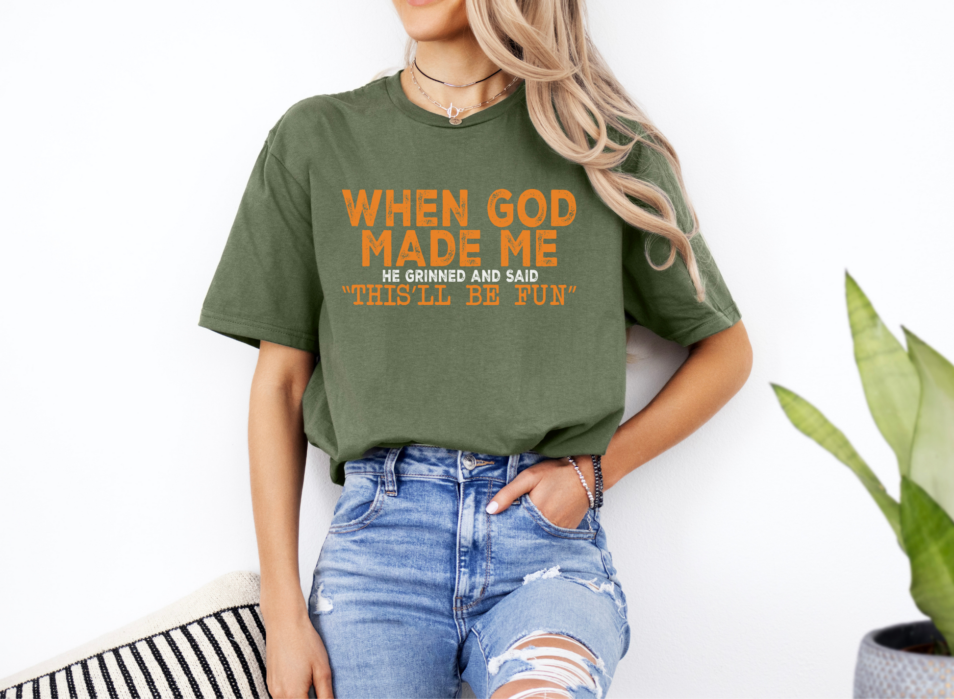 Unisex green softstyle t-shirt with orange "When God Made Me He Grinned and Said 'This'll Be Fun'" text, casual fit, paired with ripped jeans.