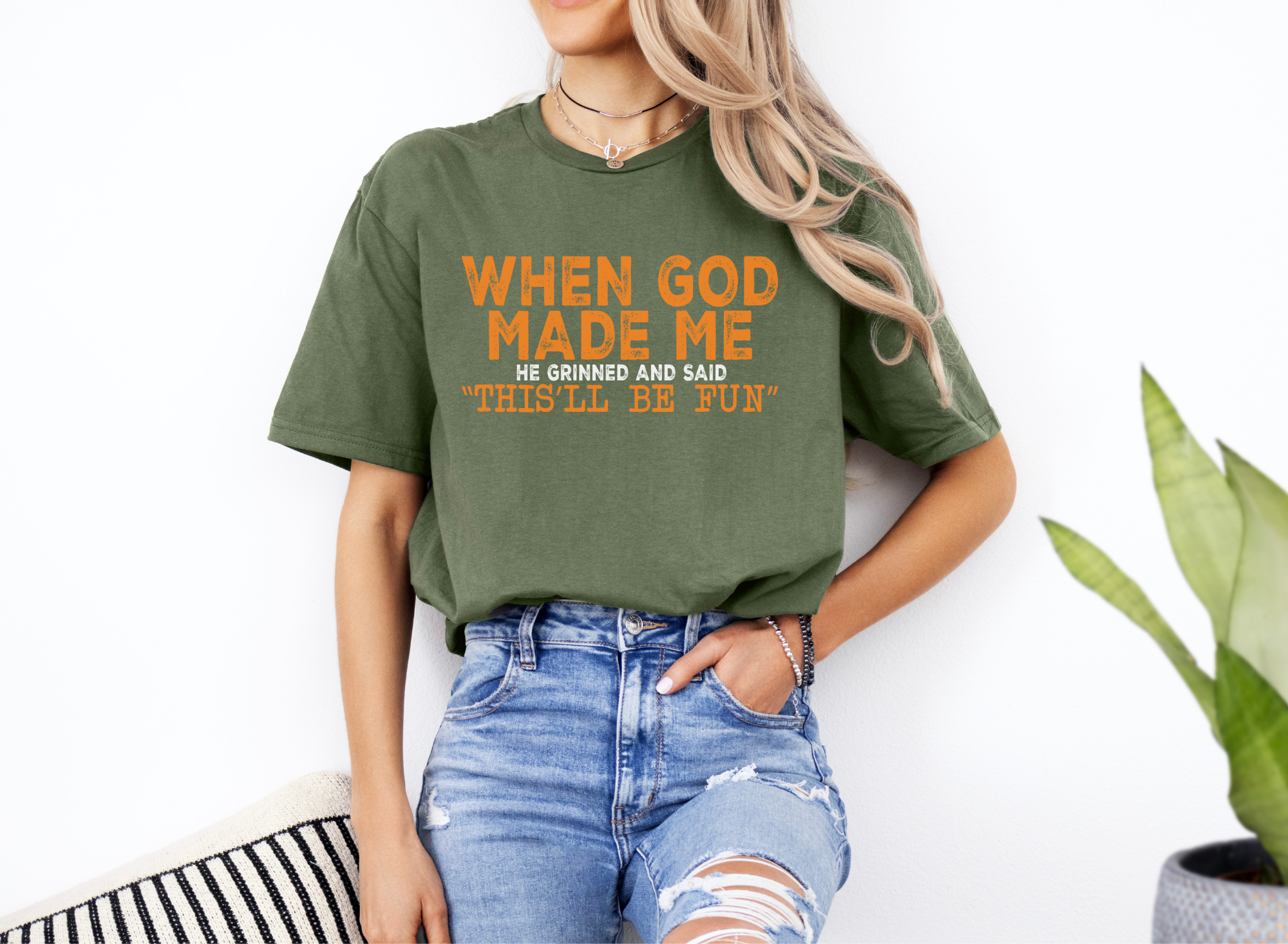 Unisex green softstyle t-shirt with orange "When God Made Me He Grinned and Said 'This'll Be Fun'" text, casual fit, paired with ripped jeans.