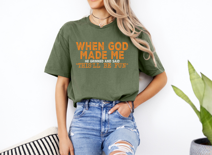 Unisex green softstyle t-shirt with orange "When God Made Me He Grinned and Said 'This'll Be Fun'" text, casual fit, paired with ripped jeans.