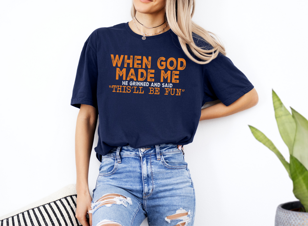 Woman wearing navy blue unisex softstyle t-shirt with orange text "When God Made Me He Grinned and Said 'This'll Be Fun'" paired with ripped jeans.