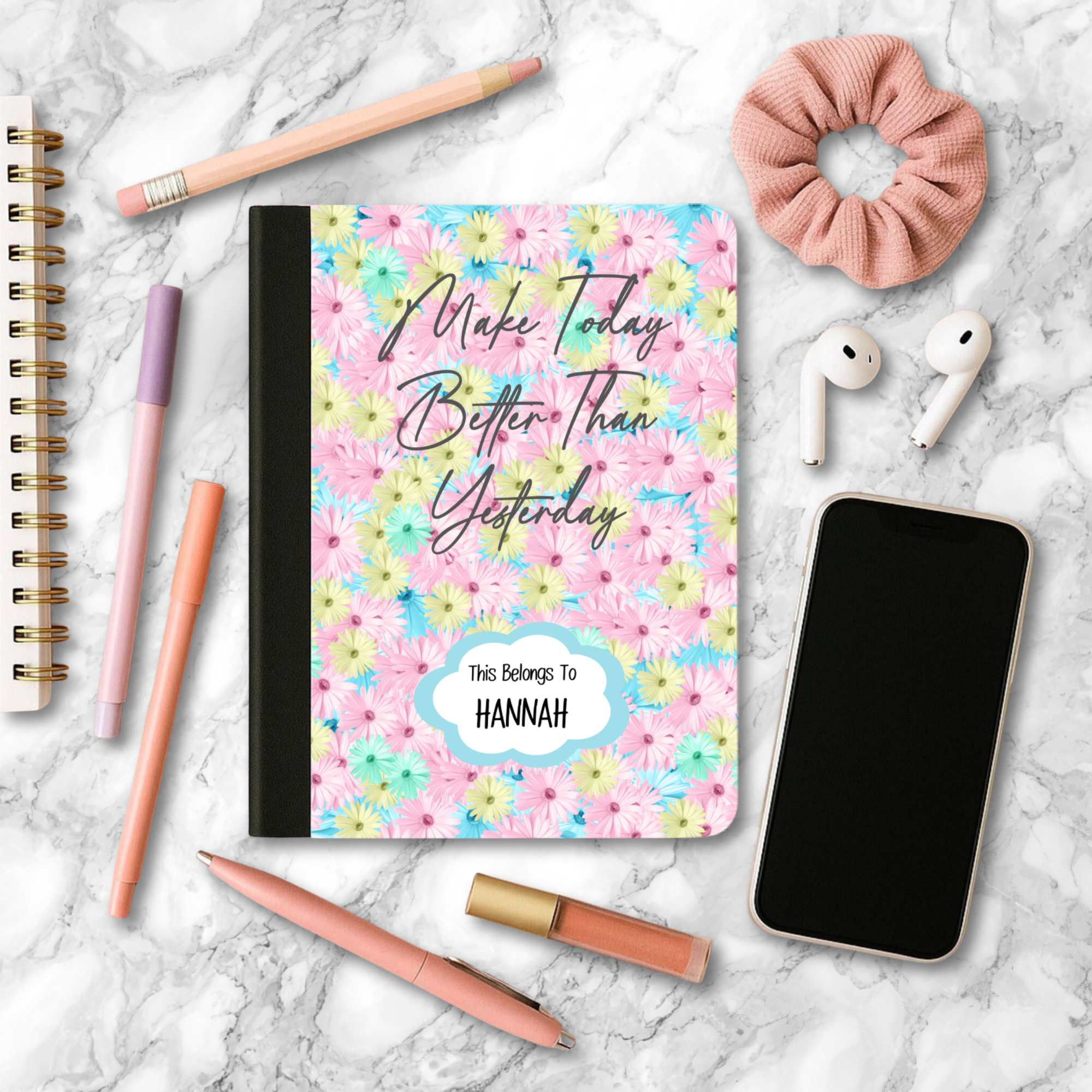 Motivational journal with pastel daisy background and personalized name "Make Today Better Than Yesterday" on cover, surrounded by pens, phone, earbuds, and scrunchie on marble surface