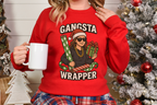 Woman wearing red Gangsta Wrapper holiday sweatshirt holding white mug by Christmas tree