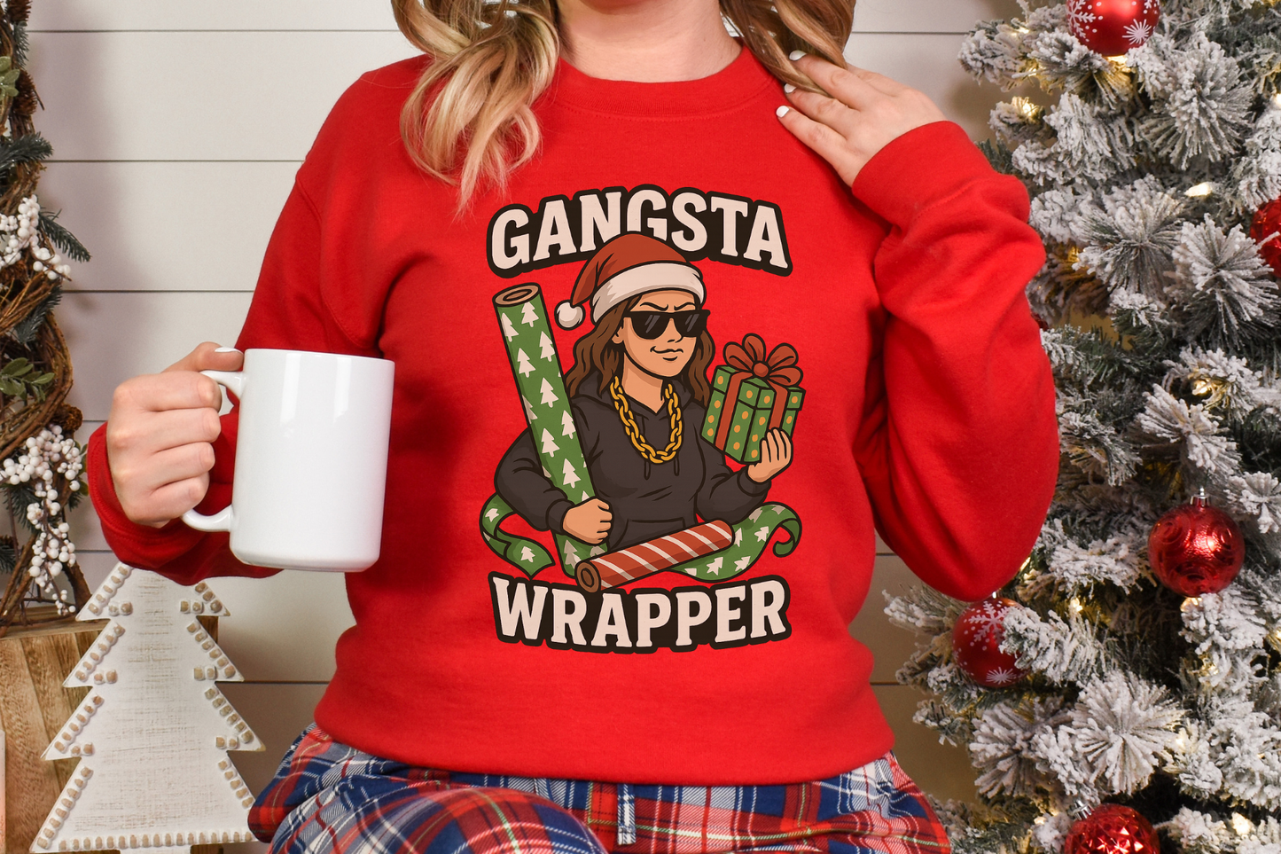 Woman wearing red Gangsta Wrapper holiday sweatshirt holding white mug by Christmas tree