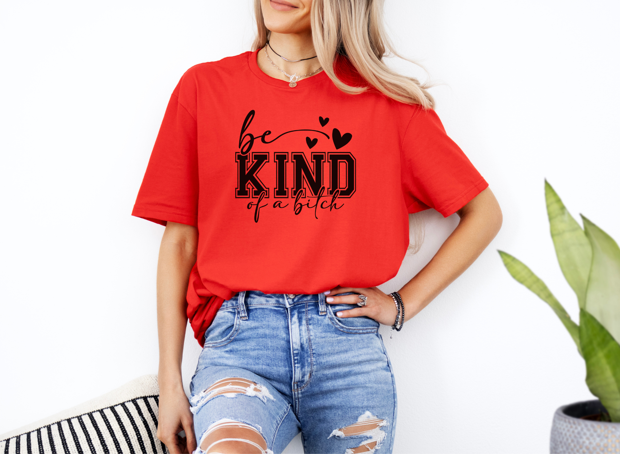 Woman wearing red "Be Kind of a Bitch" softstyle t-shirt with black bold text and hearts, paired with ripped blue jeans
