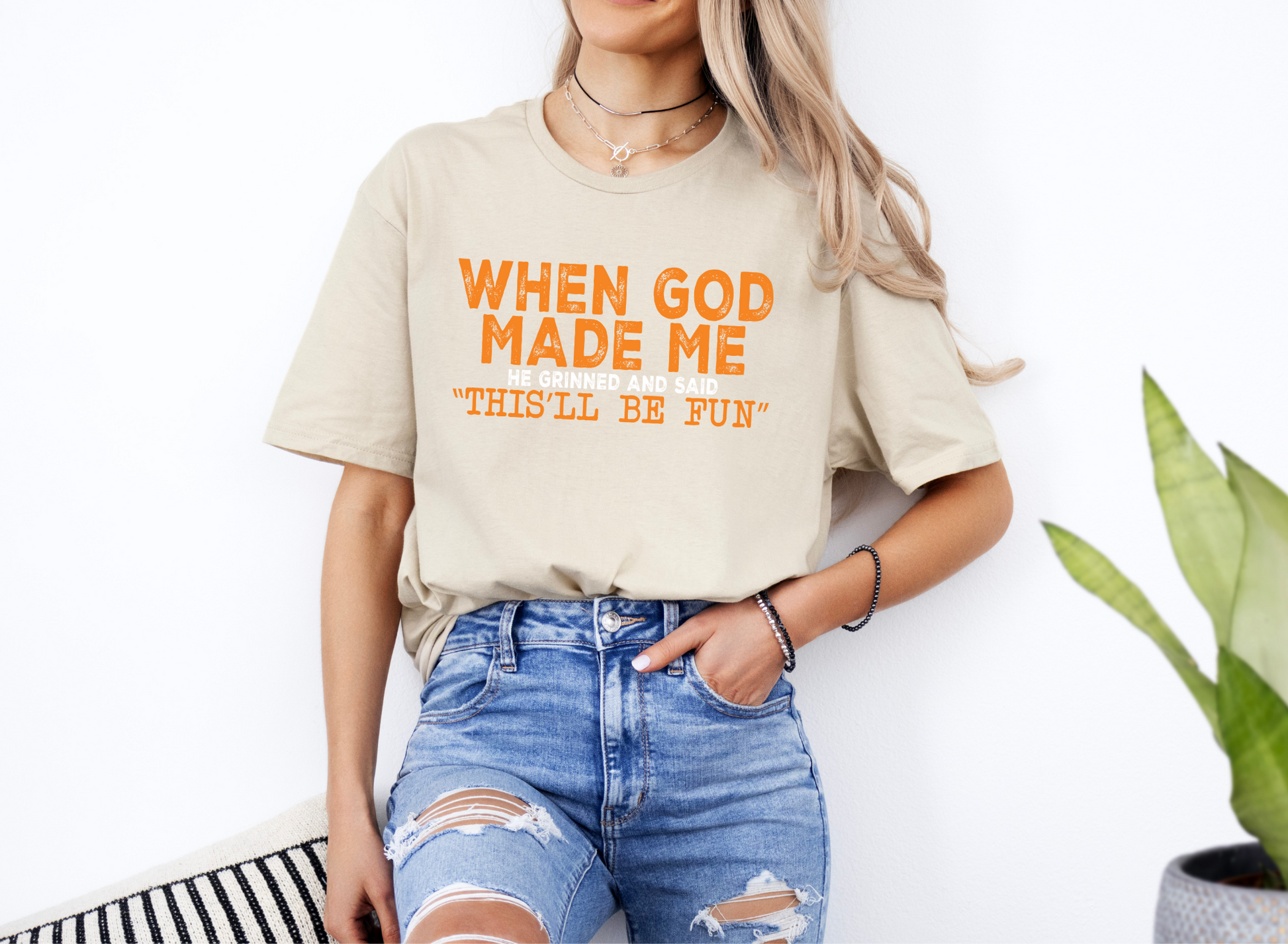 Unisex beige softstyle t-shirt with orange "When God Made Me He Grinned and Said 'This'll Be Fun'" text, styled with ripped jeans.
