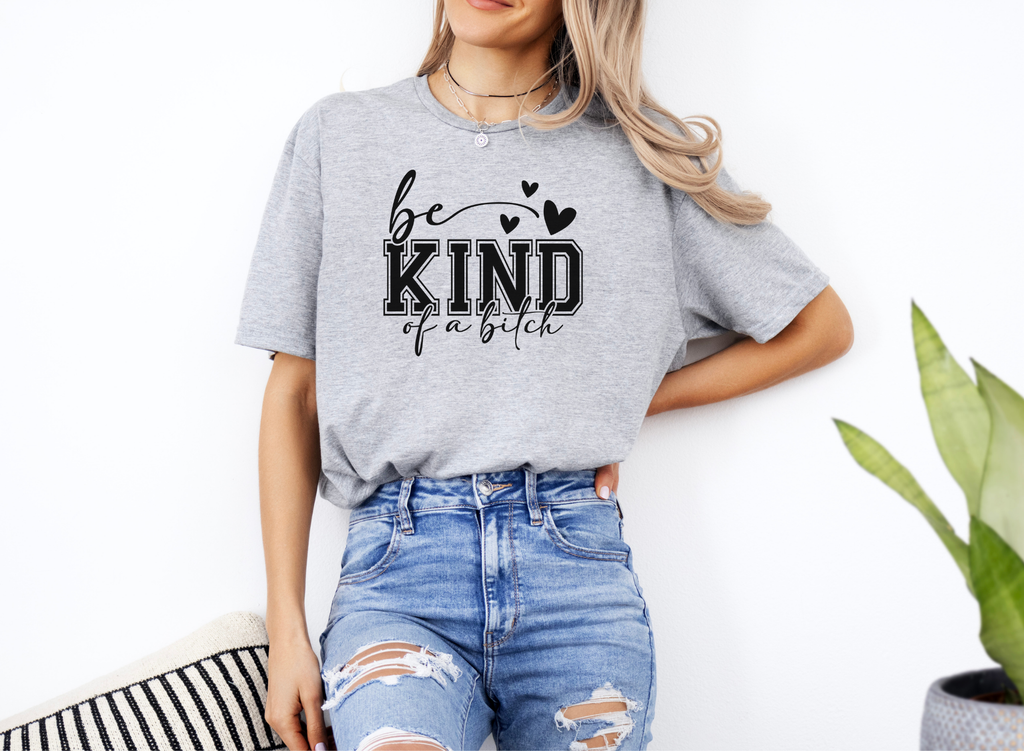 Woman wearing a grey softstyle t-shirt with bold black "be KIND of a bitch" text, styled with ripped blue jeans.