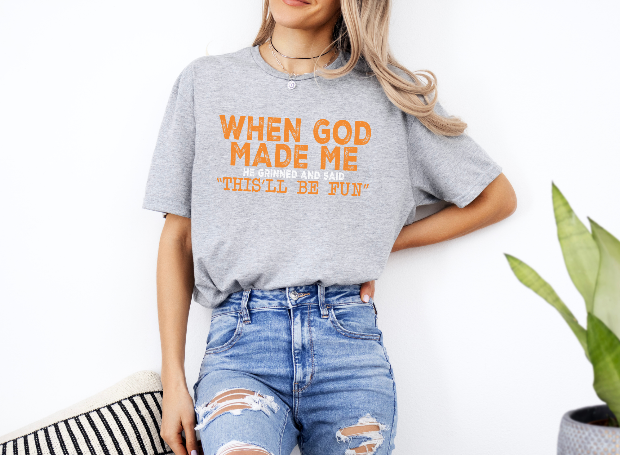 Unisex softstyle grey t-shirt with orange text "When God Made Me He Grinned and Said 'This'll Be Fun'" paired with ripped blue jeans.