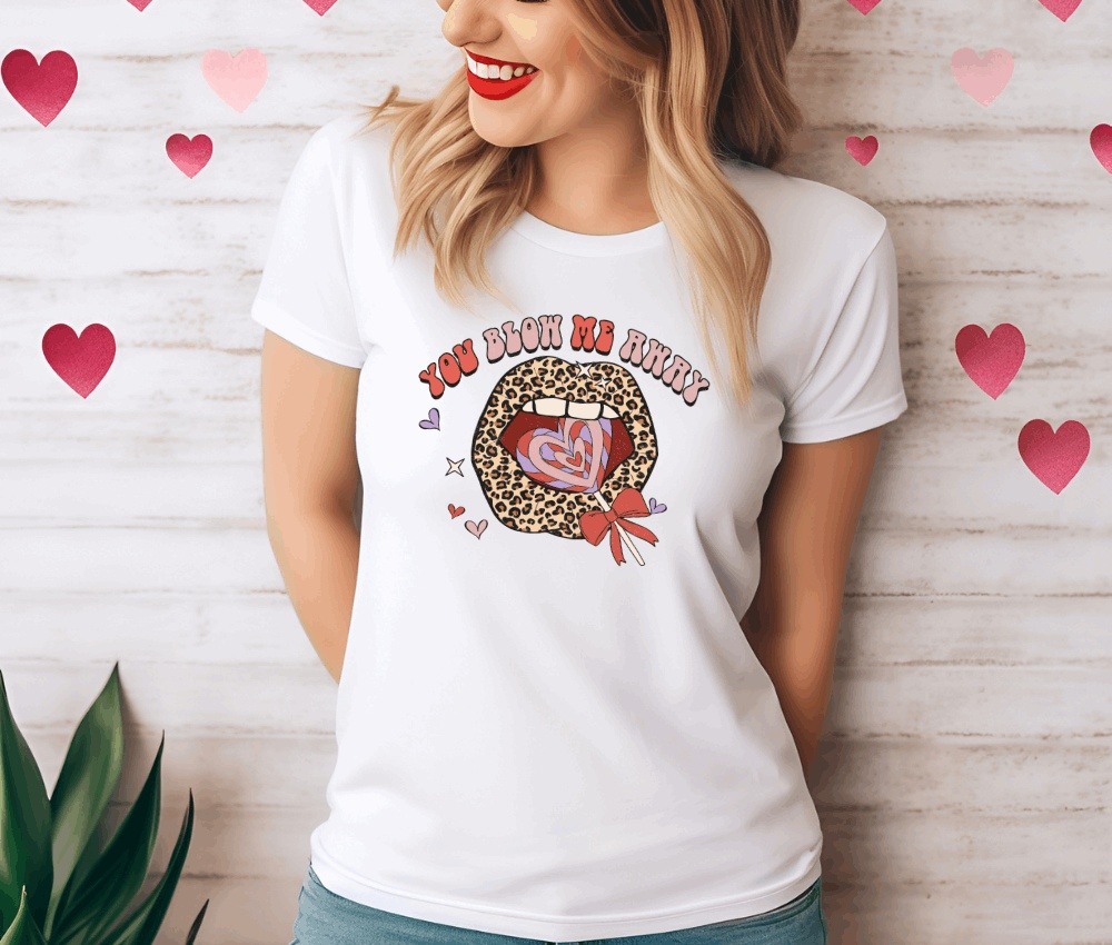 Valentine's Day t-shirt "You Blow Me Away" worn by smiling woman with heart backdrop.