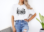 Woman wearing white "Be Kind of a Bitch" softstyle t-shirt paired with ripped blue jeans standing indoors next to a plant and cushion